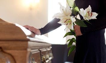 A person wearing black holds white lilies, touching a wooden casket in a softly lit room, suggesting a funeral or memorial service.
