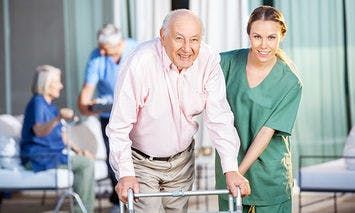 An elderly man using a walker is assisted by a smiling healthcare worker in green scrubs, with other elderly individuals and staff in the background in an outdoor courtyard setting.