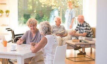 Two elderly women are sitting and talking at a table in a bright, spacious room. In the background, more elderly people interact with a caregiver near a large window.